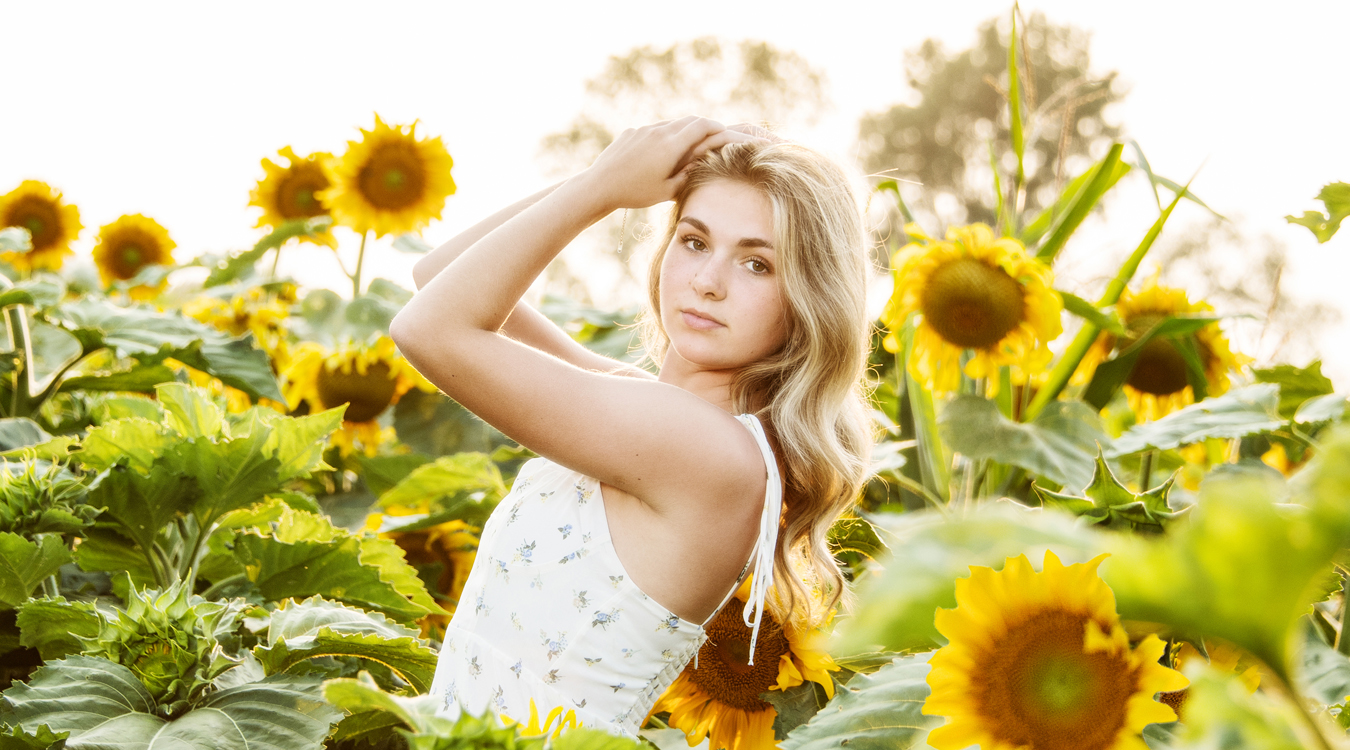 High school senior in a white floral dress standing in a backlit field of sunflowers